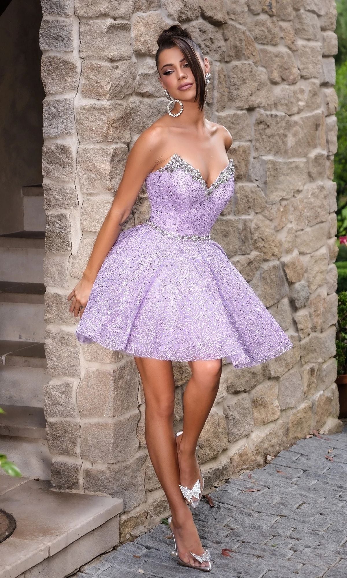 A woman stands against a stone wall in the Portia and Scarlett PS24765, a strapless sparkly lavender fit-and-flare mini homecoming dress with crystal embellishments, one leg crossed and head tilted.